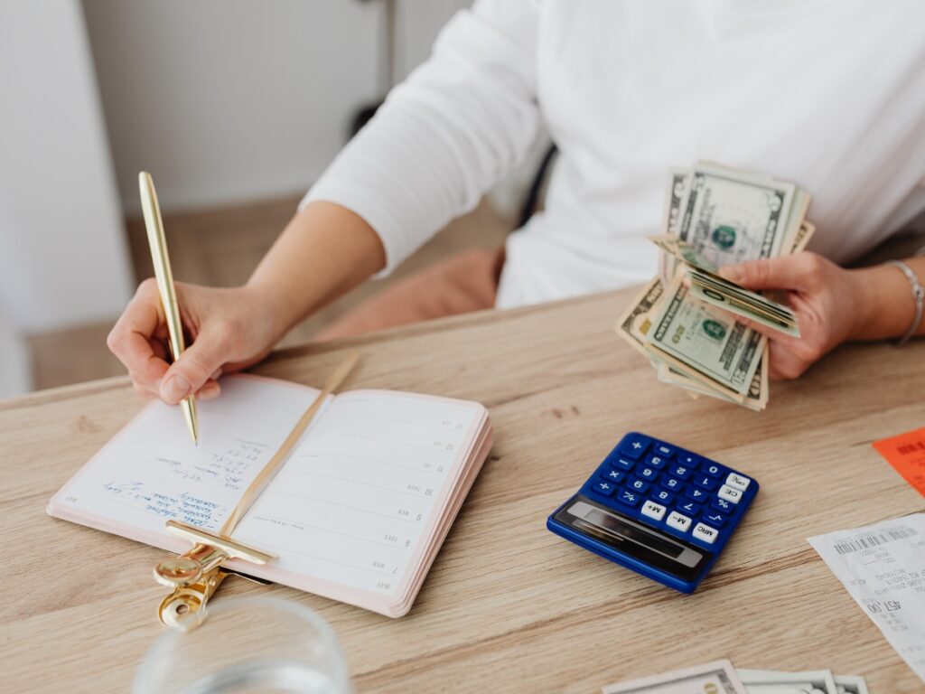 Person holding dollar bills and writing in a planner at a desk with a blue calculator and receipts, perhaps budgeting for the paver patio cost in Georgia.