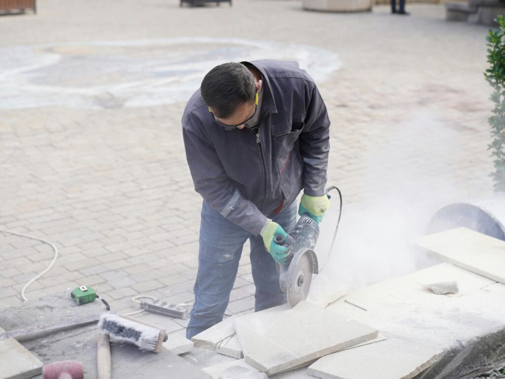 A person wearing gloves and safety glasses cuts a stone slab with an electric saw, generating dust, on a paved outdoor surface—an essential step when considering paver patio cost in Georgia.
