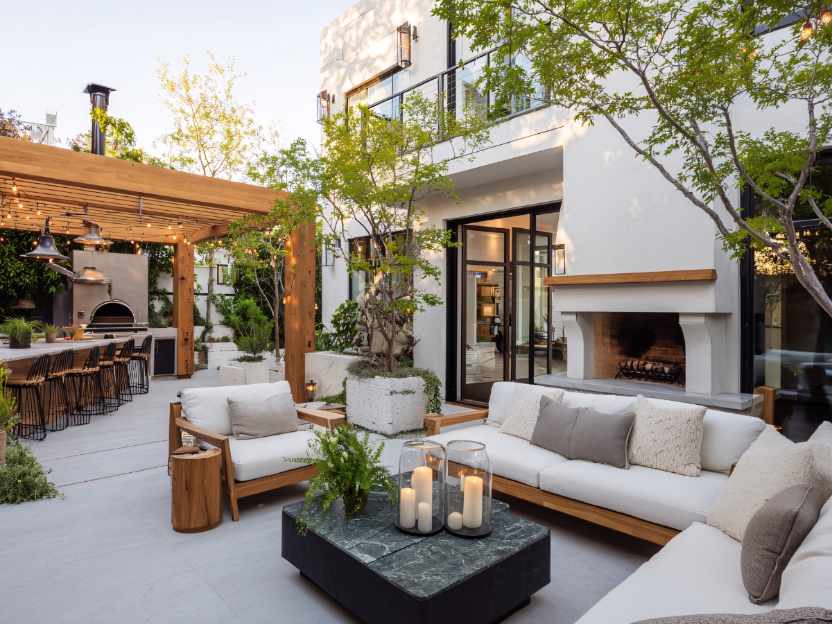 Modern outdoor patio with white sofas, a marble coffee table with candles, pergola-covered dining area, fireplace, and lush greenery—a stunning retreat that showcases the style achievable for the average paver patio cost in Georgia.