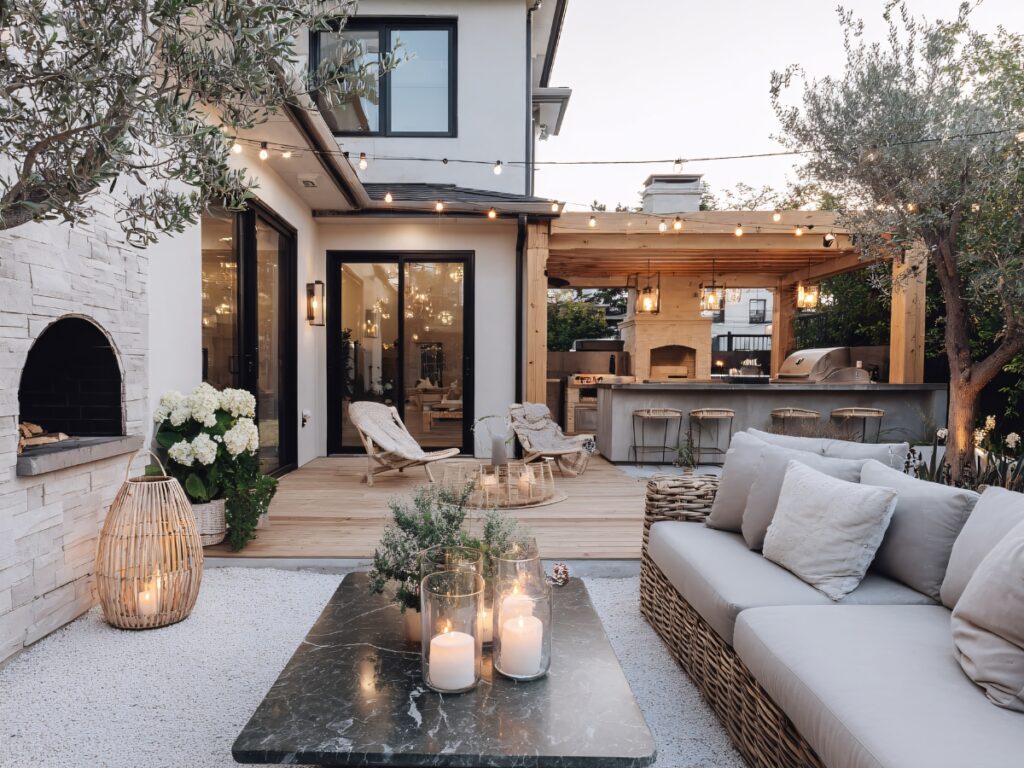 Modern outdoor patio with string lights, a sofa, a marble coffee table with candles, a wooden deck, lounge chairs, and an outdoor kitchen area with bar seating creates an inviting outdoor space for relaxing and entertaining.