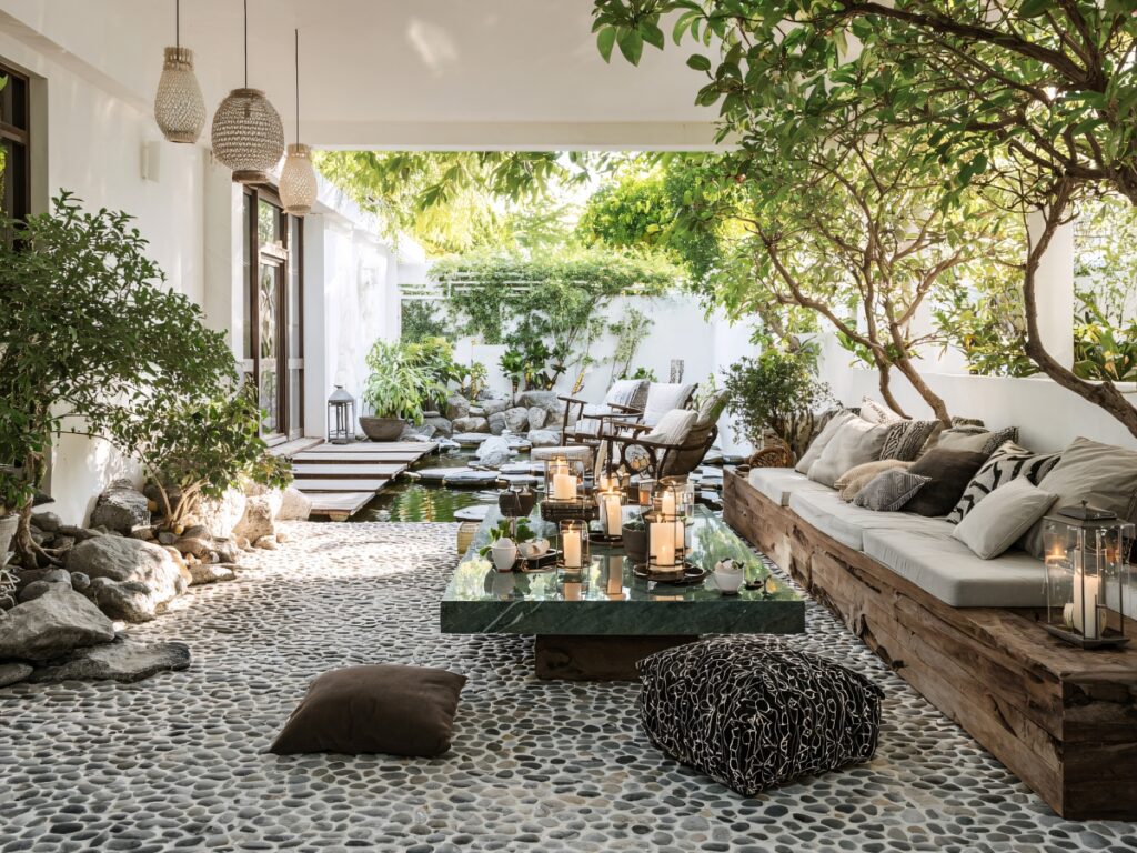 A covered outdoor space with a stone paver floor, wooden seating, a marble table with candles, and lush greenery surrounding a small pond.