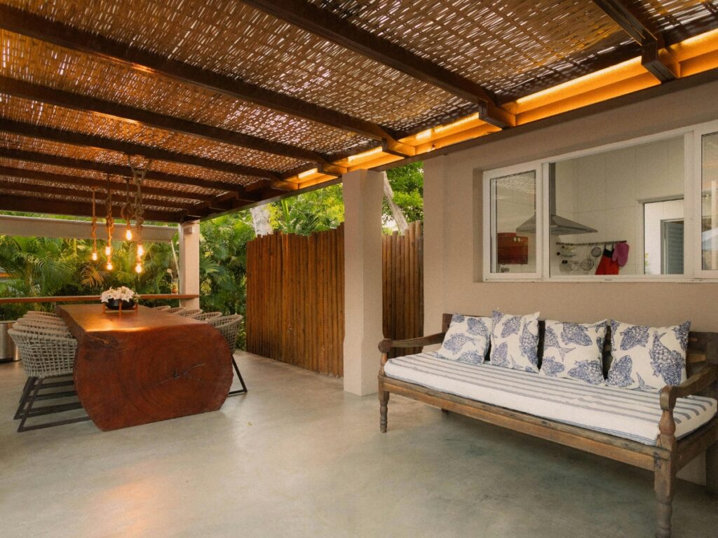 A covered outdoor space features a wooden dining table, wicker chairs, a cushioned bench with patterned pillows, and a bamboo ceiling with hanging lights over a stylish concrete patio.