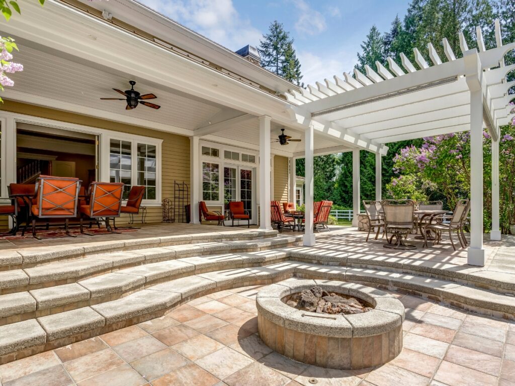Spacious outdoor space with tiered steps, a stone fire pit, dining tables and chairs on a concrete patio, a covered seating area, and a white pergola, all set against a beautiful backdrop of trees.