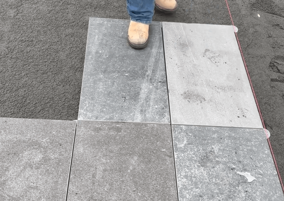 A person in work boots standing on newly laid rectangular stone tiles, with sand and a red guideline visible on the side.