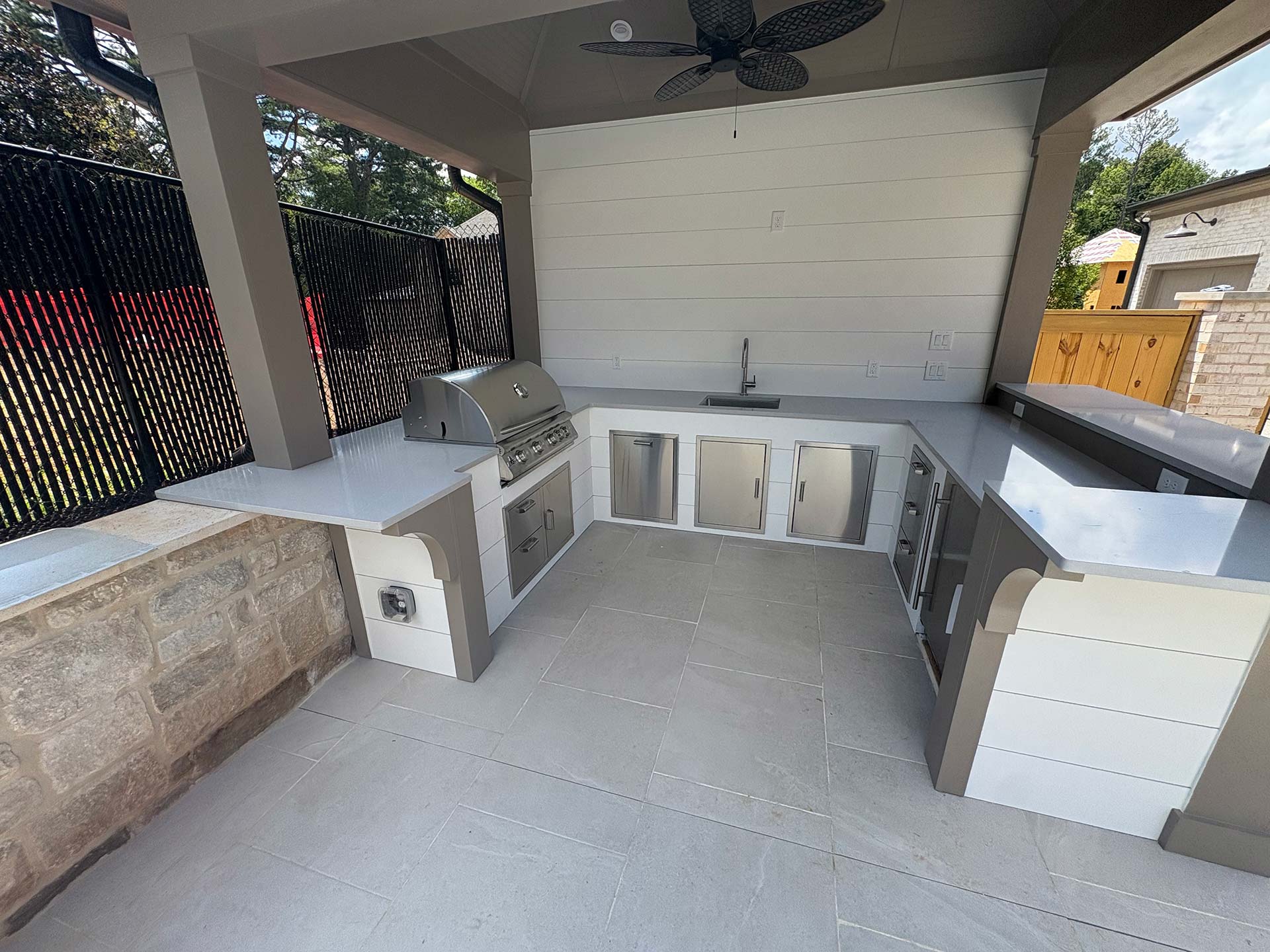 Outdoor kitchen design installation features a built-in grill, sink, cabinets, and white countertops beneath a covered patio with tile flooring.