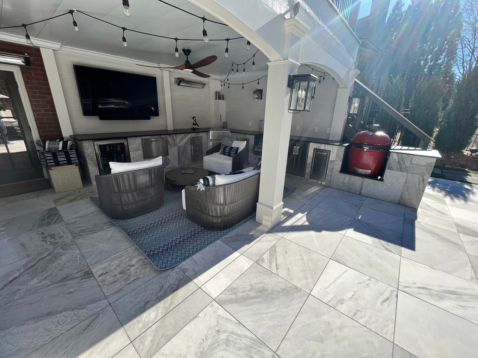 Covered outdoor patio with lounge chairs, a round coffee table on a blue rug, wall-mounted TV, ceiling fan, grill, and built-in cabinets showcasing sleek outdoor kitchen design installation on a tiled floor.