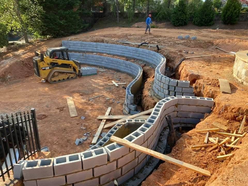 A curved cinder block retaining wall installation is underway on a dirt lot, with a skid steer loader and a person working nearby.
