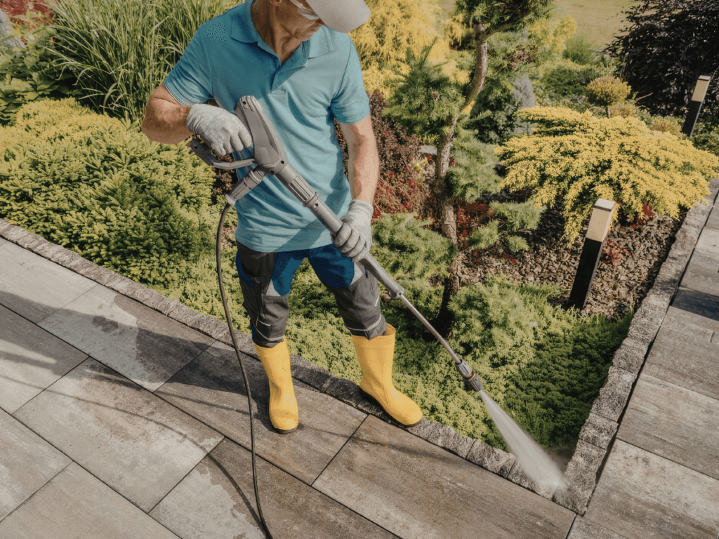 A person wearing yellow boots and gloves is pressure washing a stone patio next to a landscaped garden as part of seasonal hardscape maintenance.