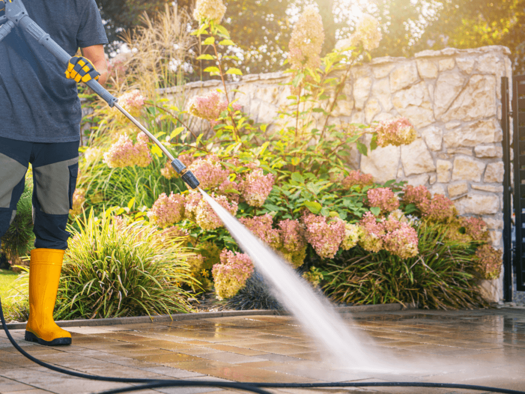 Person wearing yellow boots uses a power washer for seasonal hardscape maintenance, cleaning a stone patio in a garden with blooming hydrangea bushes and a stone wall in the background.