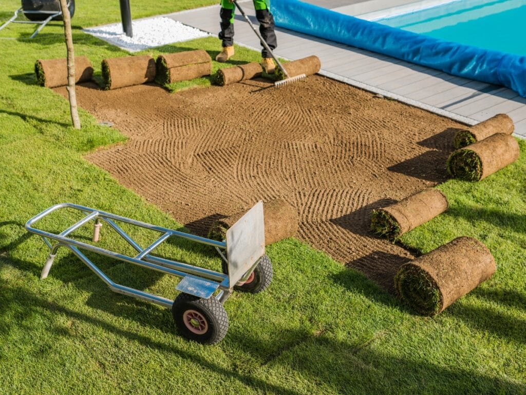 A person is raking soil to prepare for laying new sod rolls beside a swimming pool, with a wheelbarrow nearby, while considering artificial turf benefits such as low maintenance compared to traditional grass.