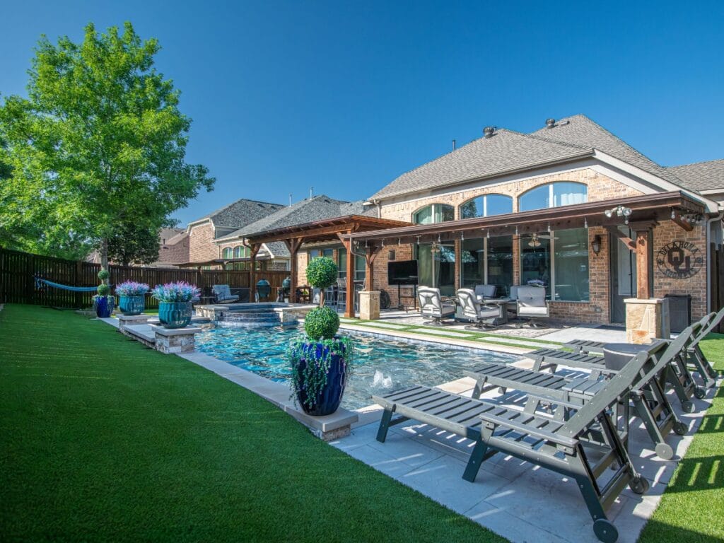 A backyard with a rectangular swimming pool, several lounge chairs set on artificial turf showcasing its benefits, potted plants, a covered patio, and brick house under a clear blue sky.