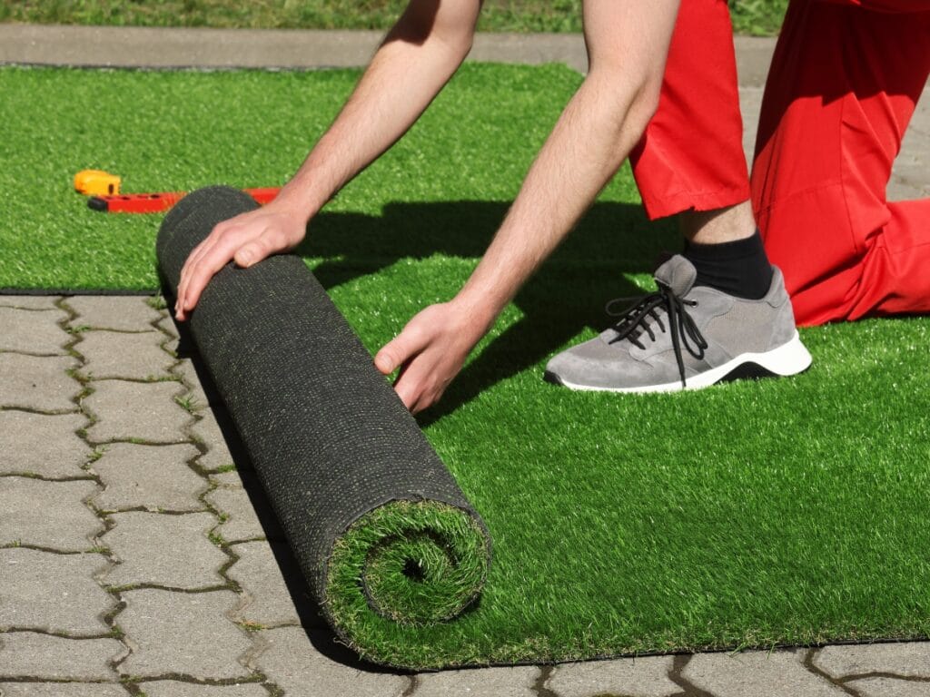 A person in red pants unrolls artificial grass onto a paved outdoor surface, with a tape measure beside them, showcasing artificial turf benefits like easy installation and year-round greenery.