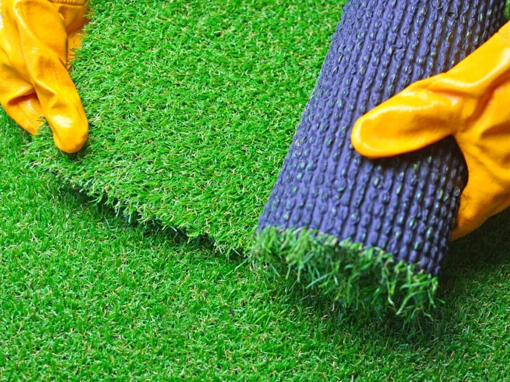 Person wearing orange gloves installing a roll of artificial grass turf onto the ground, showcasing the ease and convenience of artificial turf benefits.