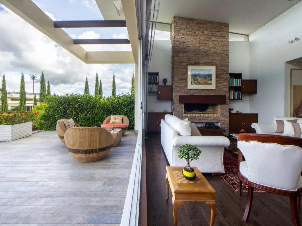 A modern living room with white sofas and a stone fireplace opens to a patio featuring paver maintenance, round wicker chairs, lush greenery, and a partly cloudy sky in the background.