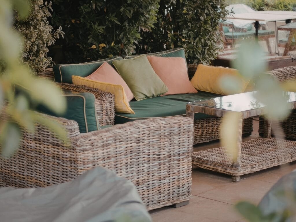 Wicker outdoor patio furniture with green cushions and colorful throw pillows surrounds a glass-top coffee table, set on well-kept pavers—thanks to regular paver maintenance—and leafy plants nearby.