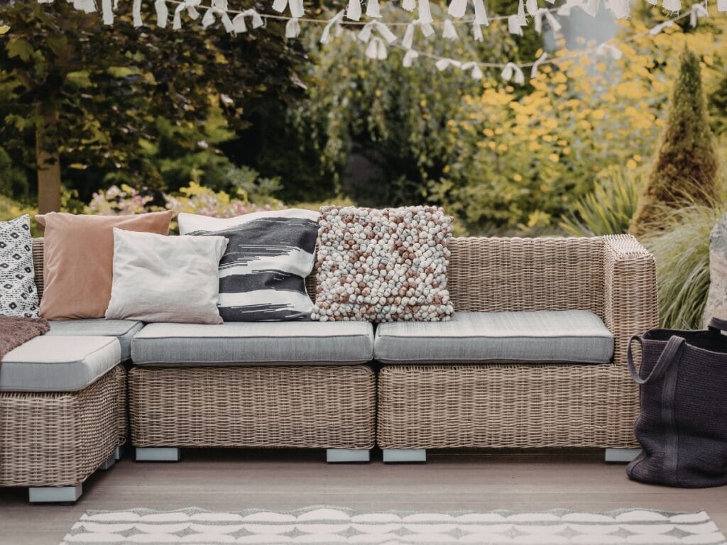 Outdoor wicker sectional sofa with gray cushions and assorted decorative pillows, set on a wooden deck surrounded by greenery and string decorations—perfect for relaxing after a day of paver maintenance.