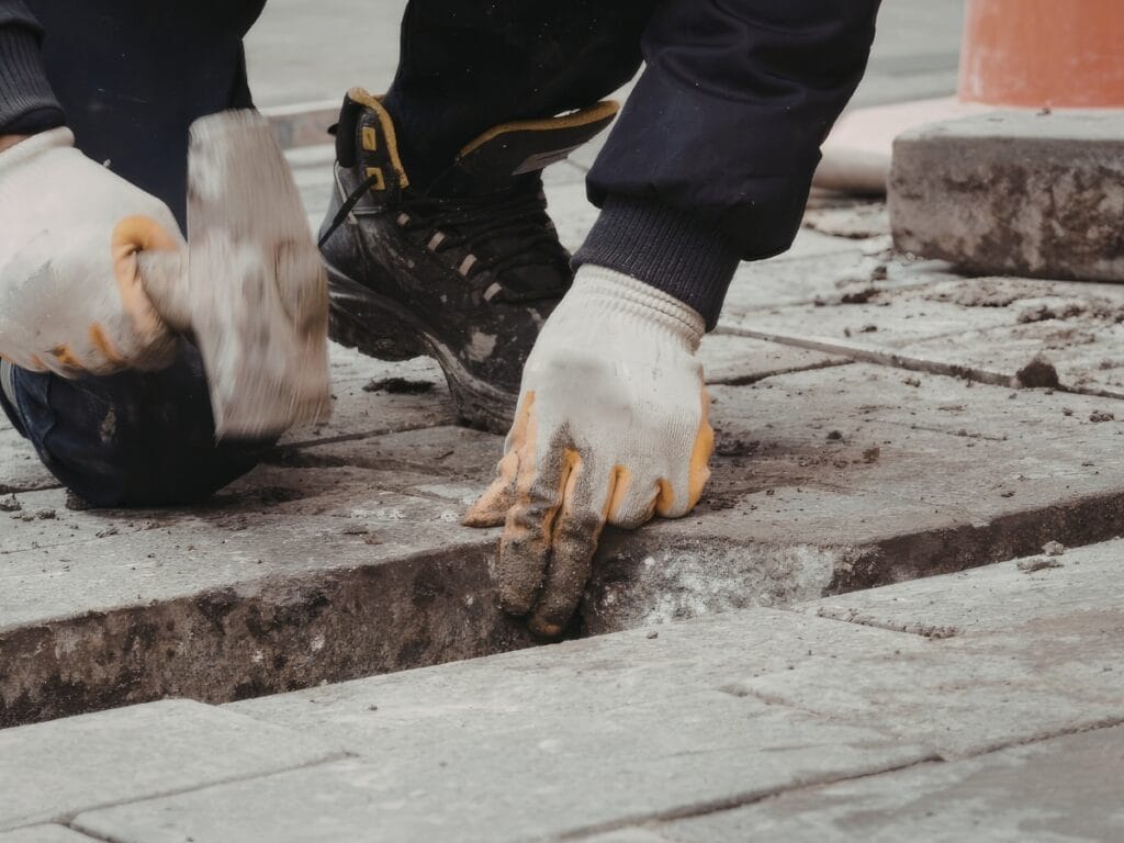 A person wearing gloves uses a hammer to install or adjust paving stones on a sidewalk, demonstrating careful paver maintenance.