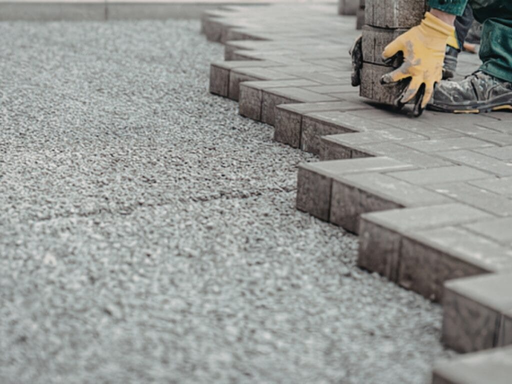 A person wearing yellow gloves and green pants is carefully placing rectangular pavers in a zigzag pattern onto a gravel surface, demonstrating expert paver maintenance.