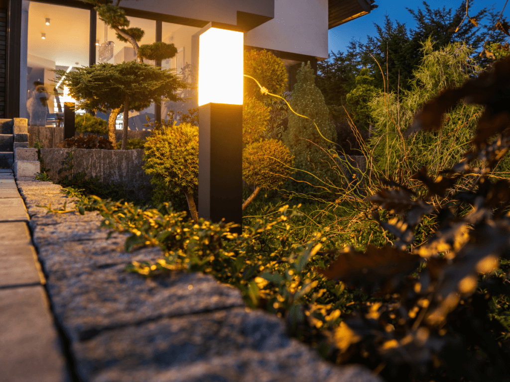 Modern outdoor pathway light, inspired by Outdoor Tech Trends, illuminates a landscaped garden at dusk, with plants and shrubs in the foreground and a person visible indoors.