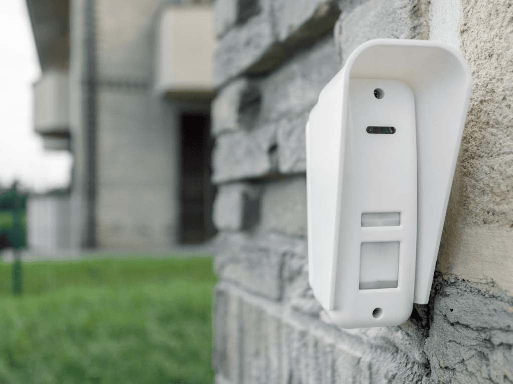 A white motion sensor device, reflecting the latest Outdoor Tech Trends, is mounted on an exterior gray stone wall with a blurred building and grass in the background.
