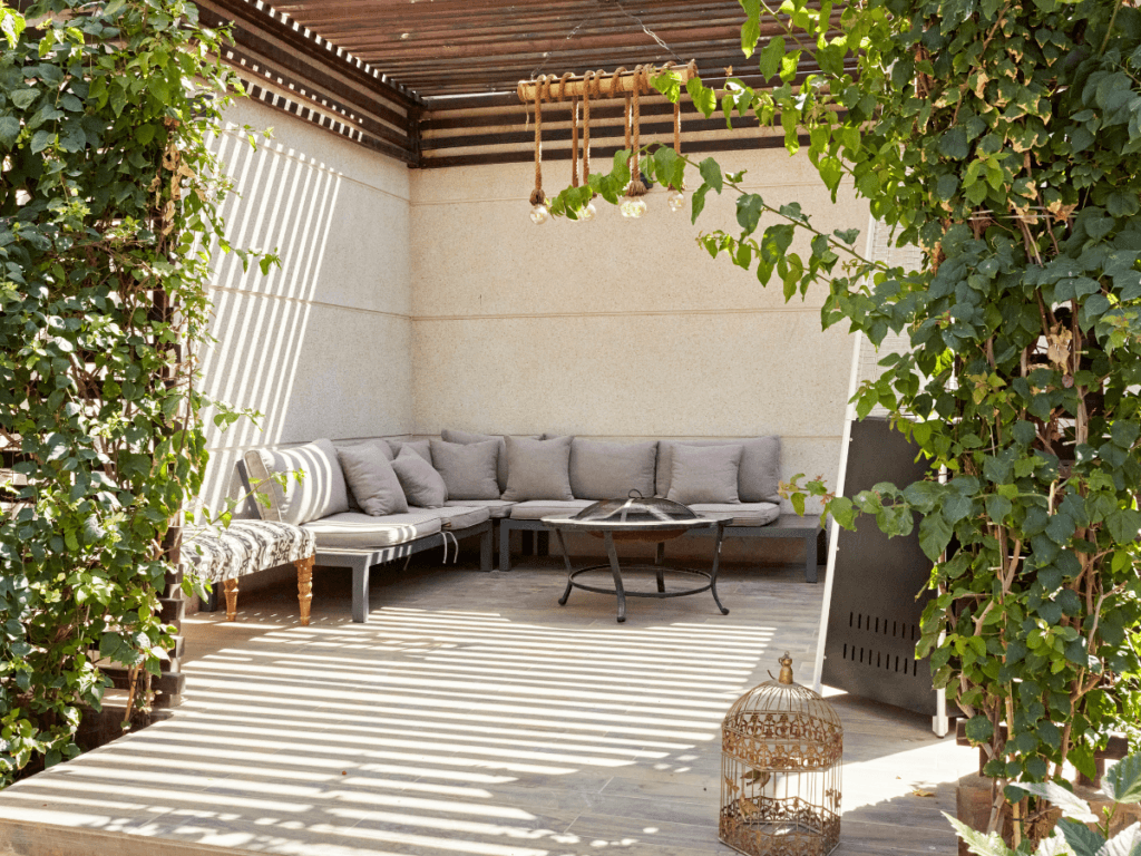 Outdoor patio with a corner sofa, round table, hanging decor, and leafy green plants partially shading the wooden deck; a decorative birdcage sits on the floor, blending comfort with the latest Outdoor Tech Trends.