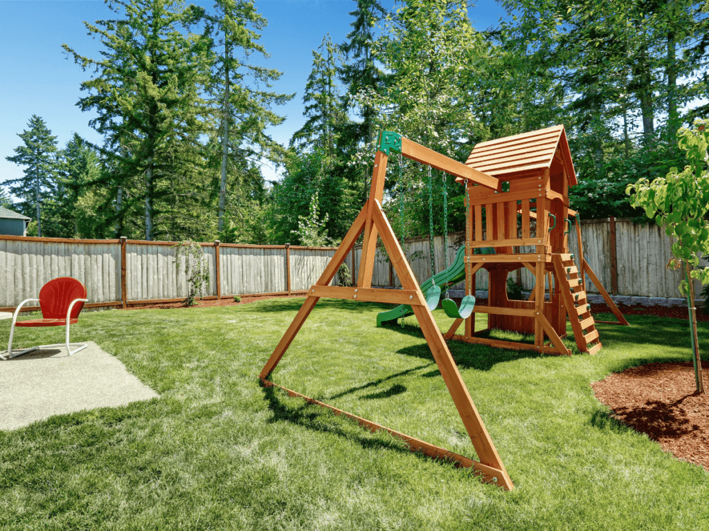 A backyard with a wooden playset including swings and a slide, designated kids play areas with a patch of sand and a red chair, all enclosed by a wooden fence surrounded by trees.