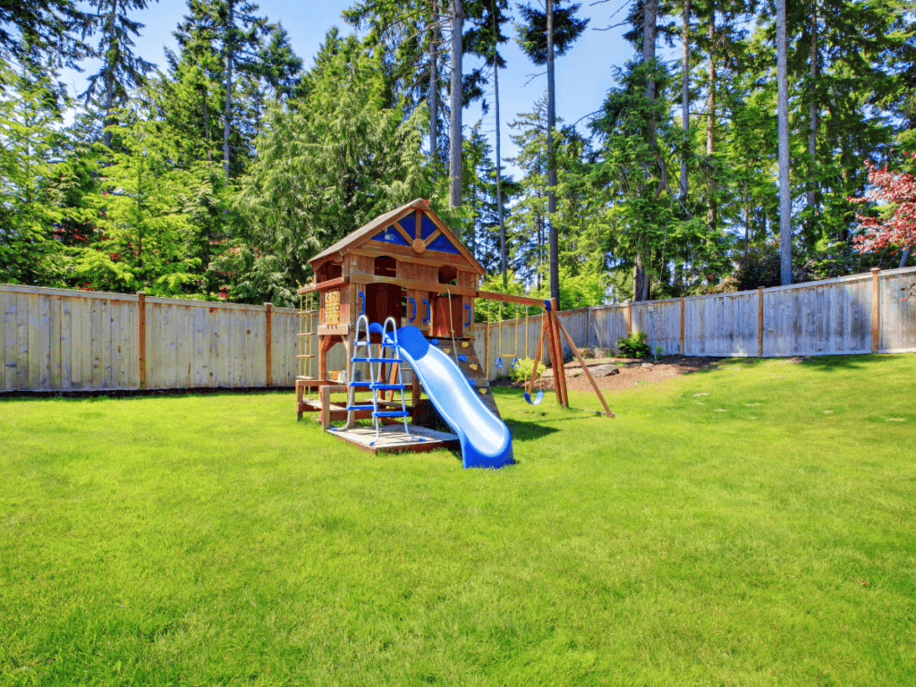 A wooden playset with a blue slide, swings, and climbing features stands on a grassy lawn surrounded by a wooden fence and tall trees—an inviting spot perfect for kids play areas.