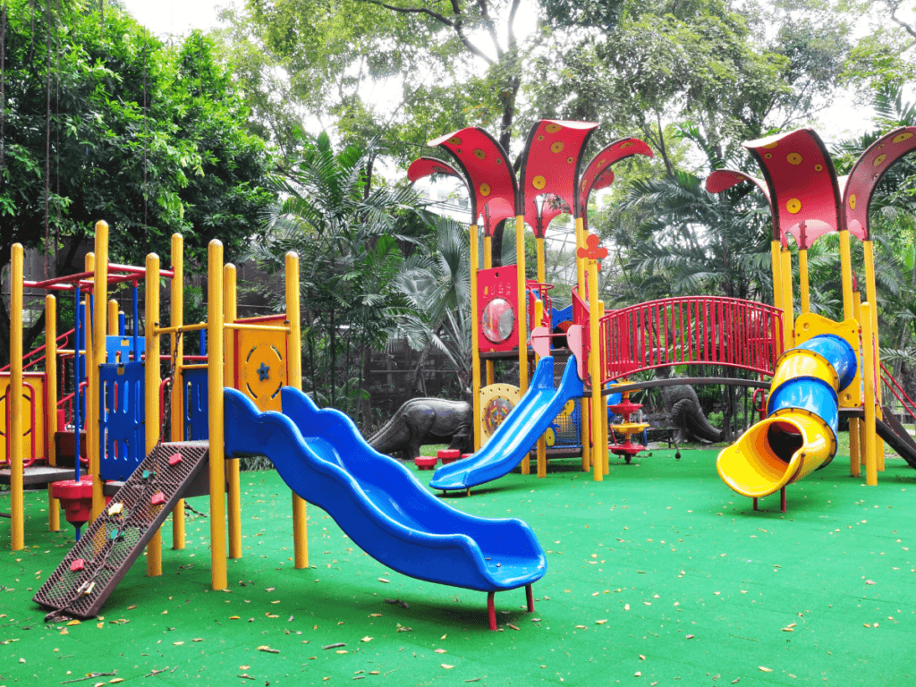 Colorful outdoor playground with slides, climbing structures, and a bridge on green artificial turf, surrounded by trees and greenery—perfect for fun-filled kids play areas.