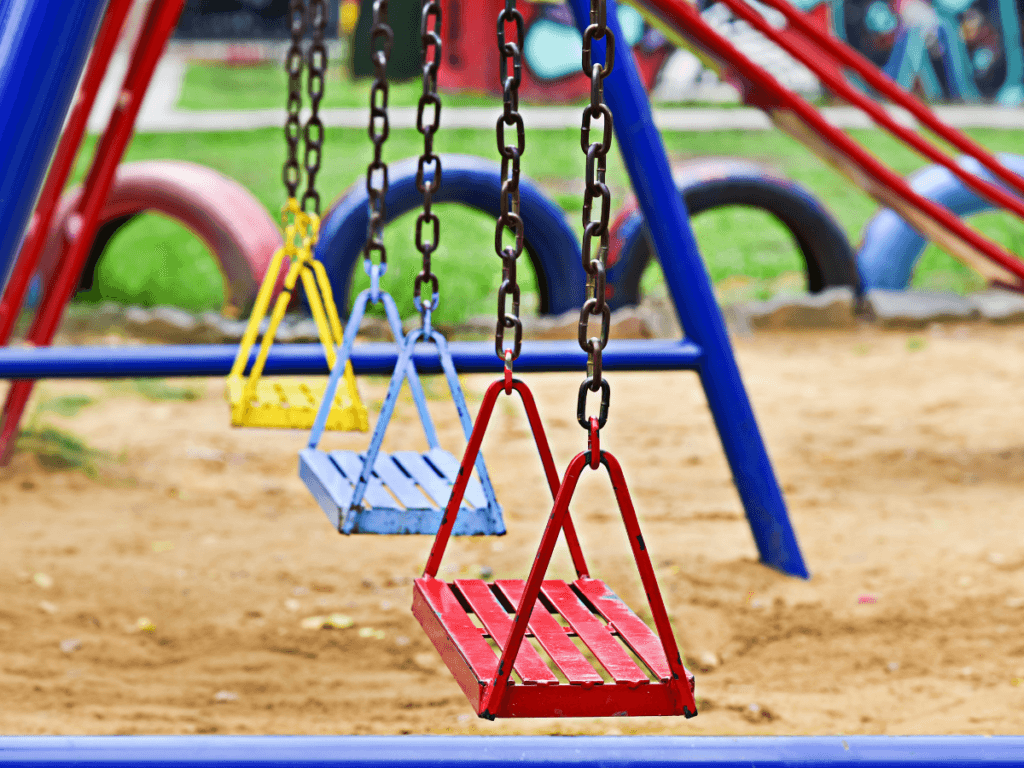 Three empty wooden swings painted red, blue, and yellow hang from chains on a playground, part of vibrant kids play areas with sand and colorful tires in the background.