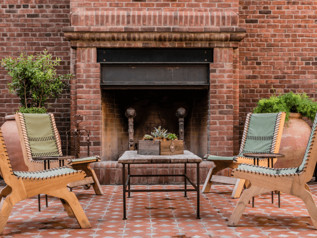 Outdoor patio with two wooden chairs, a small table featuring a plant arrangement, large potted plants, and a brick fireplace—perfect for those seeking stylish small yard solutions.