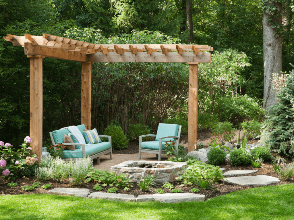 A wooden pergola with two light blue cushioned outdoor sofas sits on a stone patio, offering stylish small yard solutions amid lush greenery and a landscaped garden.