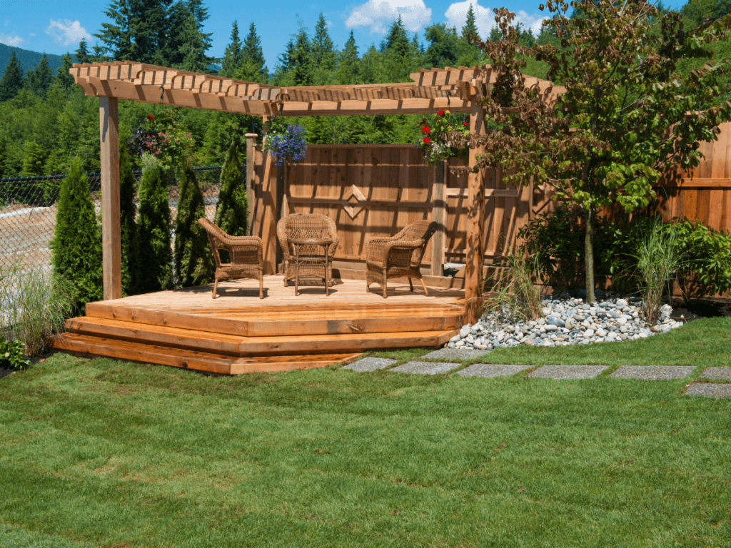 A wooden pergola with wicker chairs and a table sits on a raised deck in a landscaped backyard, offering stylish small yard solutions with green grass and a stone path.