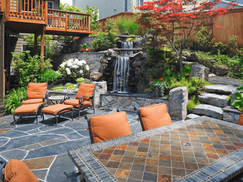 Backyard patio with orange cushioned chairs, a tiled table, stone flooring, and a tiered rock waterfall surrounded by plants and a Japanese maple tree—an inviting example of stylish small yard solutions.