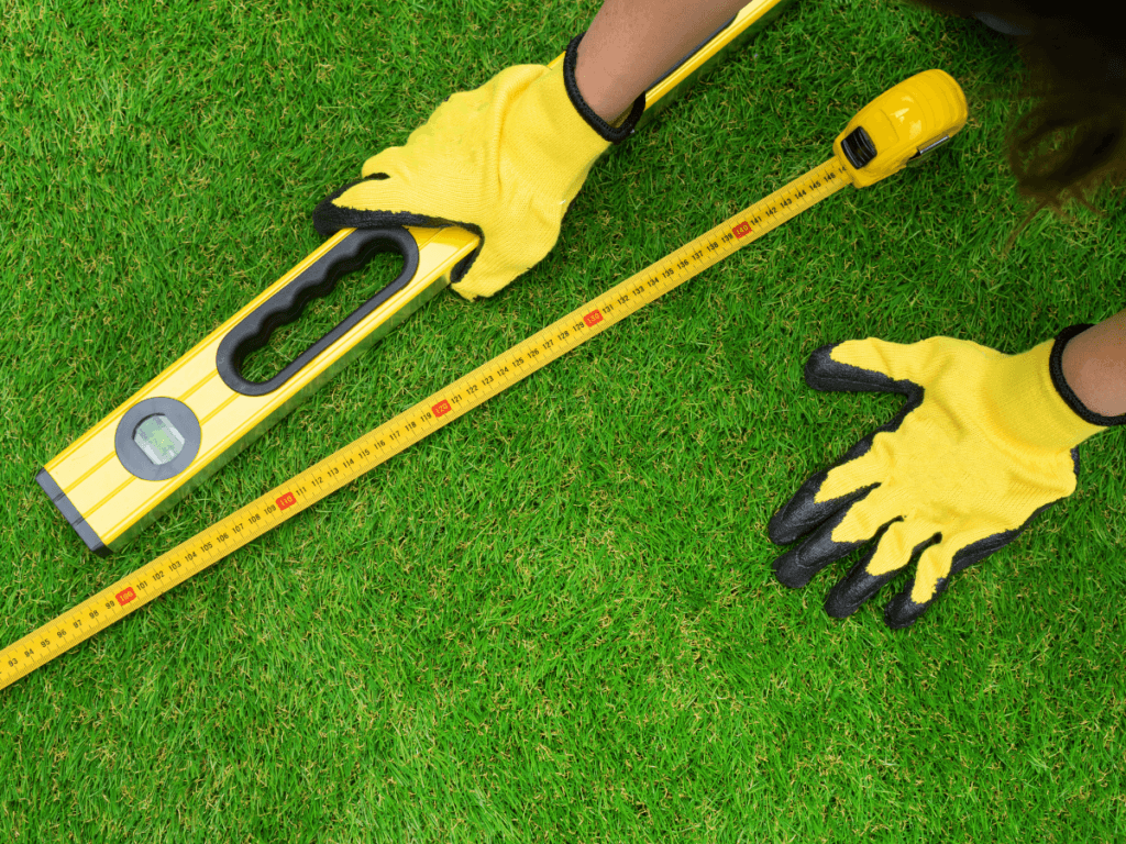 Person wearing yellow gloves measures artificial turf with a yellow tape measure and holds a bubble level tool.