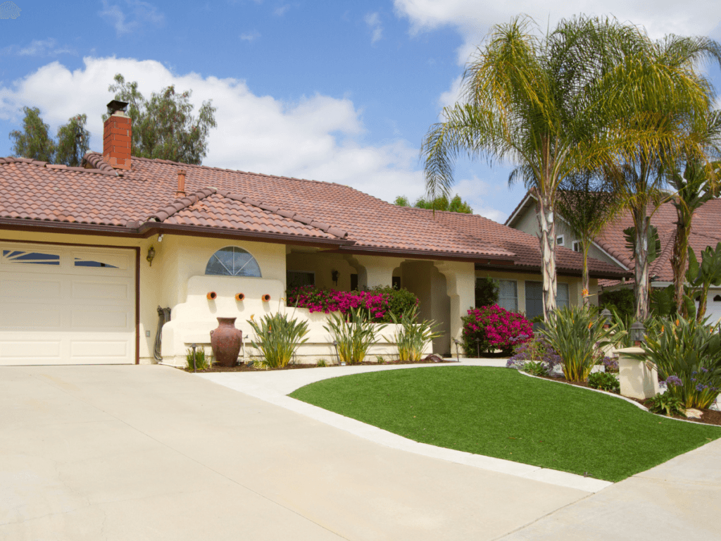 Single-story house with a red tile roof, large driveway, lush artificial turf lawn, palm tree, and flowering shrubs under a blue sky with scattered clouds.
