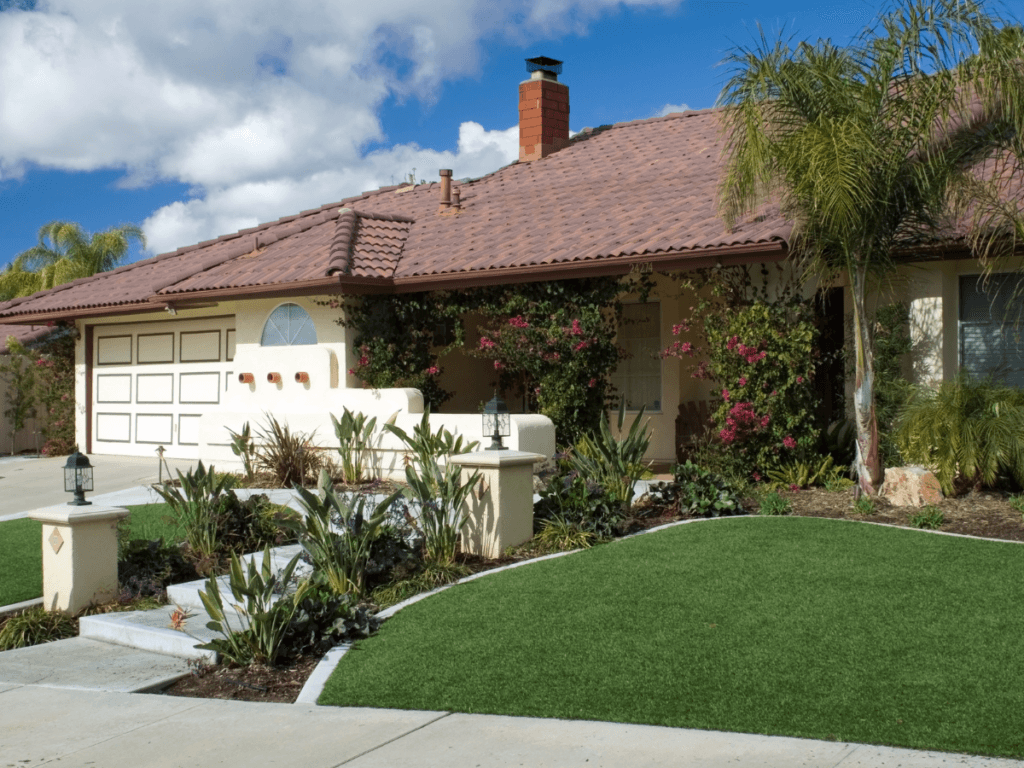 A single-story house with a tile roof, attached garage, lush landscaping, palm trees, and a manicured artificial turf lawn under a partly cloudy sky.