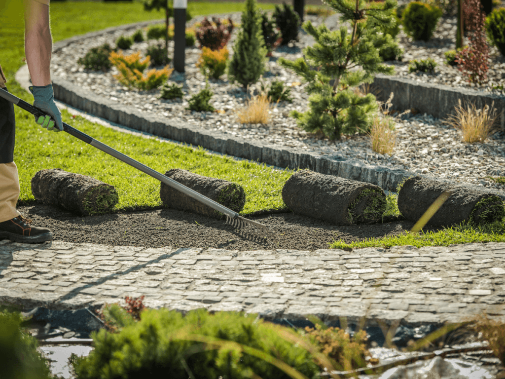 A person levels soil with a rake next to rolled-up sod pieces and artificial turf in a landscaped garden with stone paths and various plants.