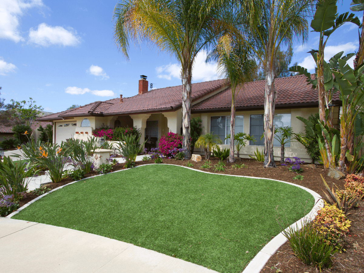 Single-story house with a red tile roof, surrounded by palm trees, tropical plants, colorful flowers, and a neatly manicured green lawn of artificial turf under a partly cloudy sky.