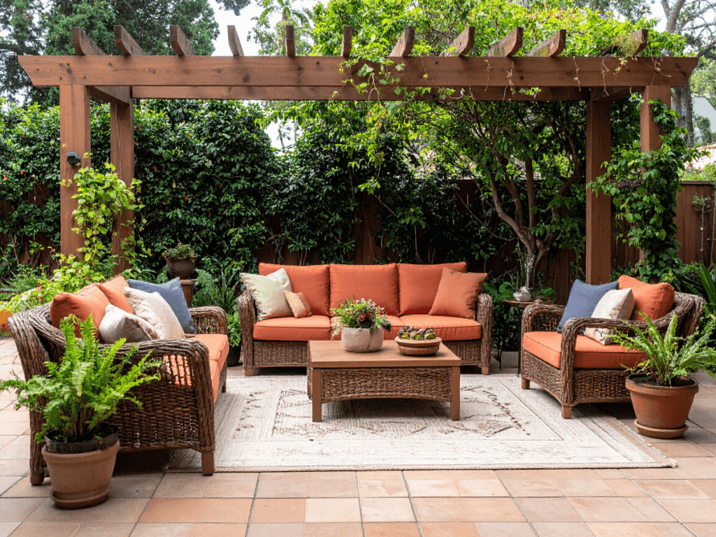 Outdoor patio with wicker furniture, orange cushions, a wooden coffee table, potted plants, rug, and a wooden pergola surrounded by greenery—offering inviting shade solutions for relaxing in comfort all day long.