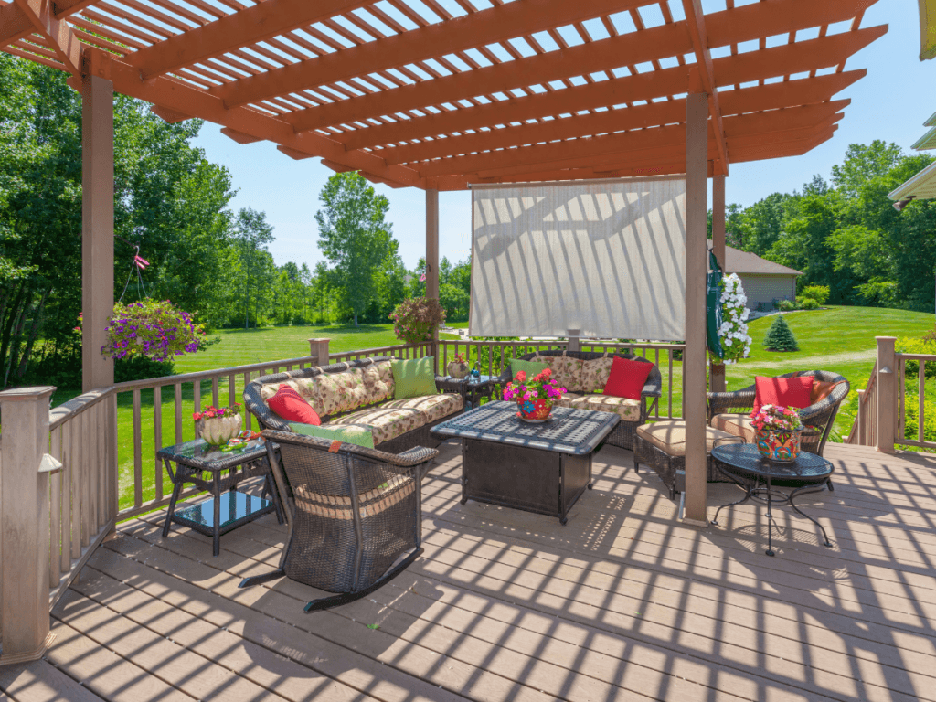 A wooden pergola provides elegant shade solutions for an outdoor patio with cushioned seating, a fire pit table, flowers, and a view of a green lawn and trees.