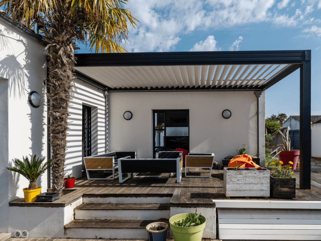 Outdoor patio with modern pergola offering intelligent shade solutions, complemented by black and wood furniture, various potted plants, and a palm tree, attached to a white house under a partly cloudy sky.