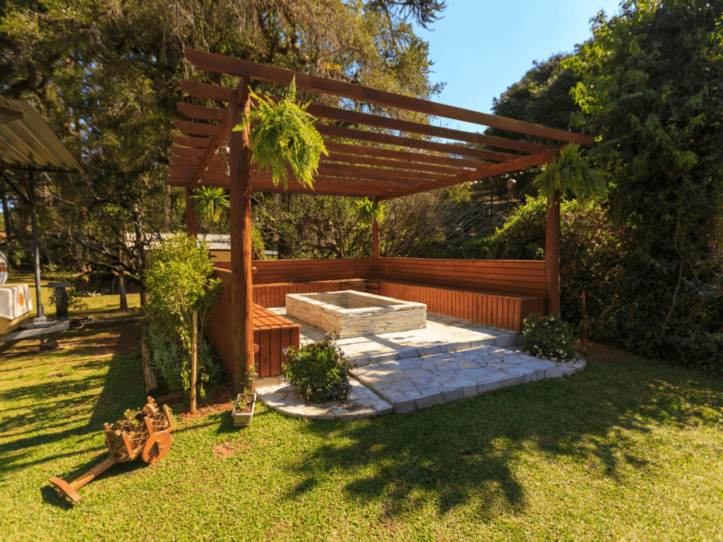 Wooden pergola with potted plants, a bench, and a raised stone planter on a paved patio—one of the most attractive shade solutions—set in a grassy backyard with trees and shrubs.