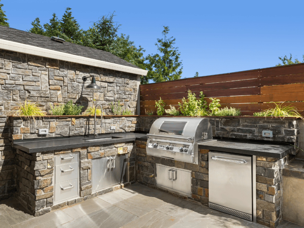 Outdoor kitchen with built-in stainless steel grill, sink, fridge, and other premium outdoor kitchen appliances, featuring stone countertops and herb planters set against a stone wall and wood fence, with trees and blue sky in the background.