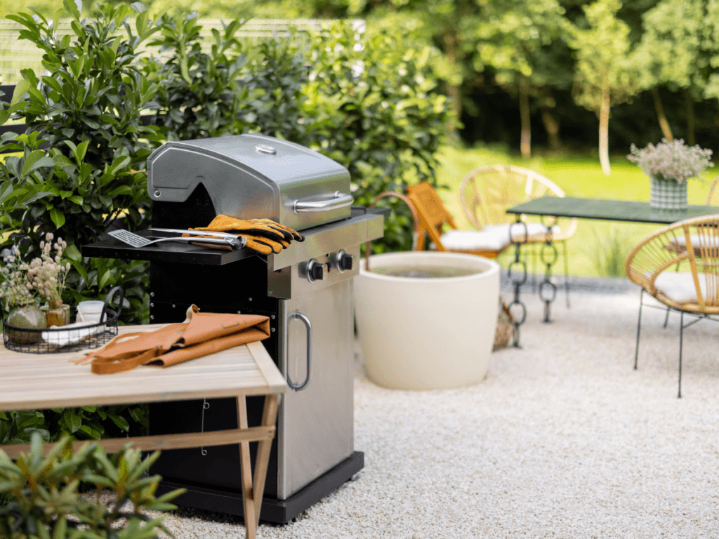 A stainless steel gas grill, one of the must-have outdoor kitchen appliances, with gloves and utensils sits on a patio near a table and chairs, surrounded by greenery and outdoor plants on a sunny day.
