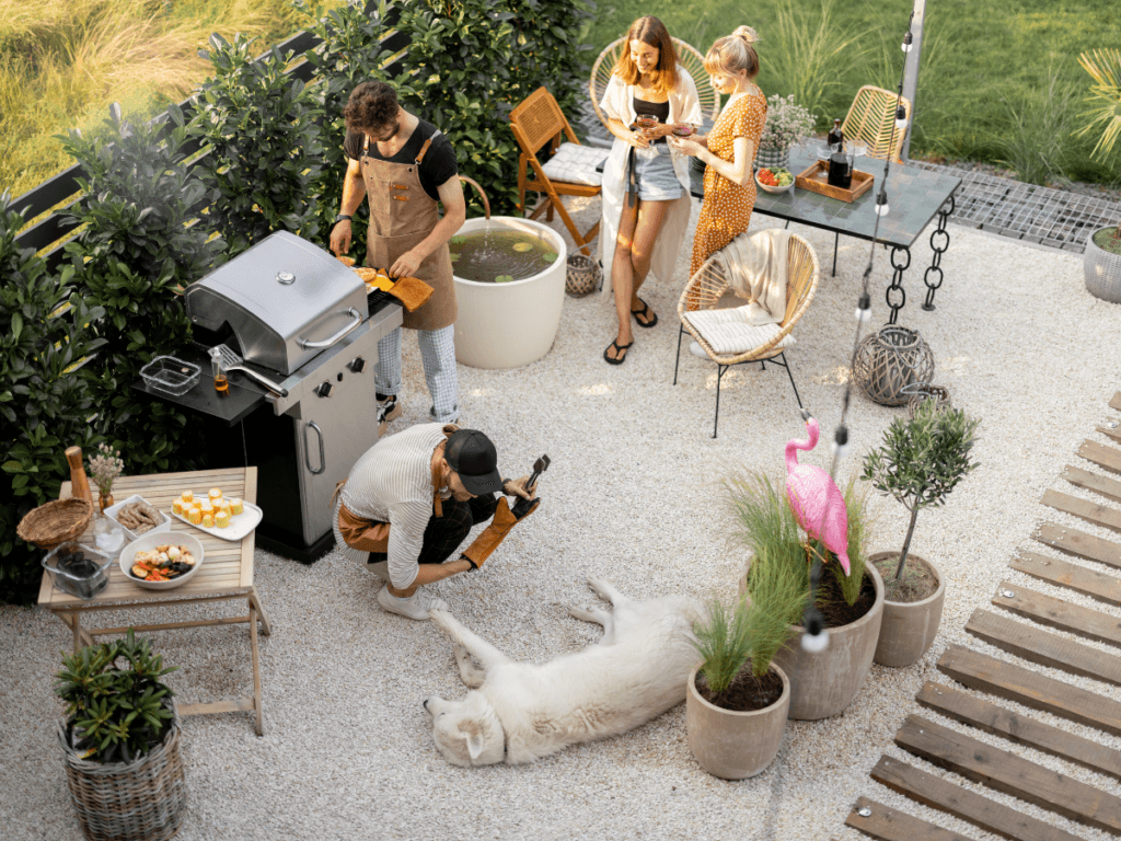 Four people have a backyard barbecue; one tends the grill, another pets a large white dog lying on the ground, while two women prepare food nearby using outdoor kitchen appliances. Patio plants and outdoor décor are visible.