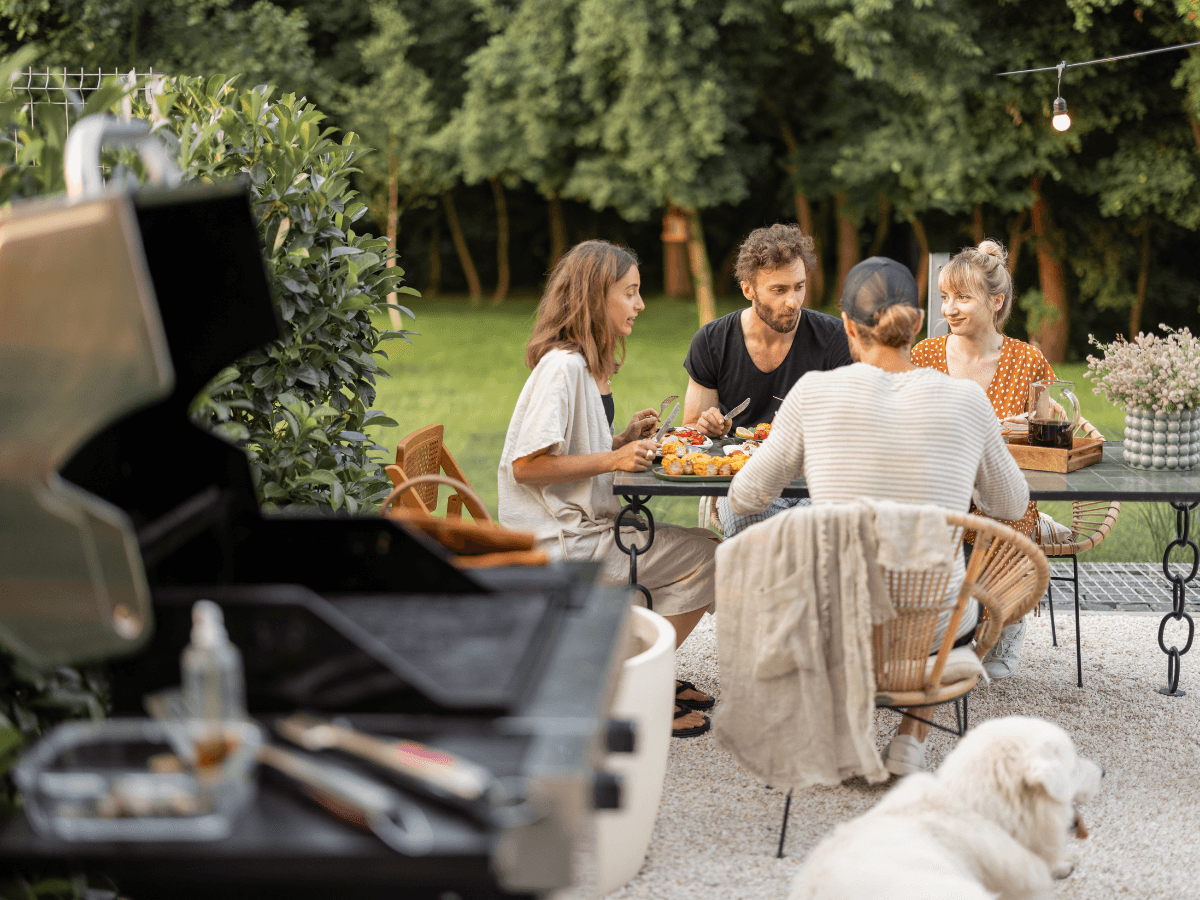 Four people sit around an outdoor table eating a meal in a garden, with outdoor kitchen appliances and a grill beside them and a white dog in the foreground. Trees and greenery are visible in the background.