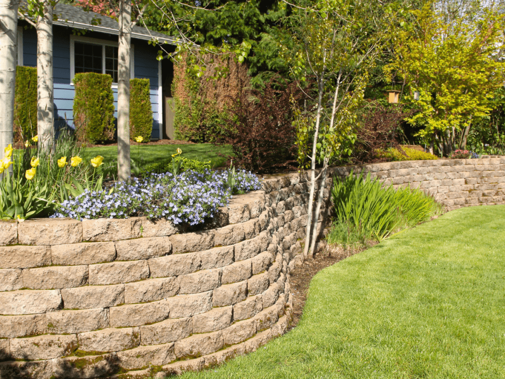 A landscaped yard with a curved segmental retaining wall, blooming flowers, green grass, and various trees in front of a house.