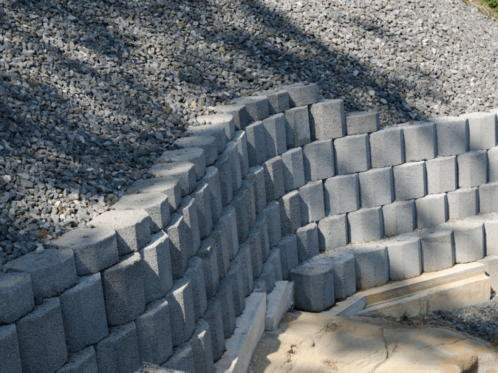 A tiered segmental retaining wall made of stacked concrete blocks sits in front of a sloped area covered with loose gravel.