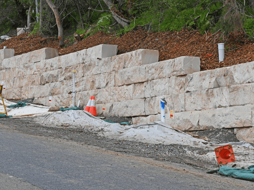 A large segmental retaining wall is under construction beside a road, with traffic cones, pipes, and construction materials scattered on the ground.